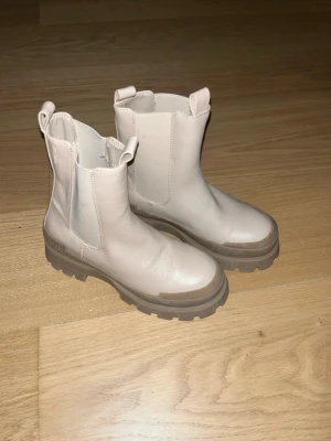 Beige chunky chelsea boots - Säljer ett par beige chelsea boots med grov sula och elastiska paneler på sidorna. Skorna har dragflikar både fram och bak för enkel påtagning. Tillverkade i skinnliknande material och har en riktigt chunky vibe.