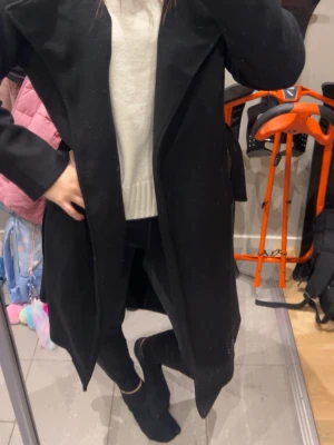 Svart lång kappa med bred krage - Säljer en svart, lång kappa med bred krage och rak passform. Jackan har en stilren look och är perfekt för dig som gillar minimalistisk stil. Passar till lager på lager och är enkel att matcha med olika outfits.