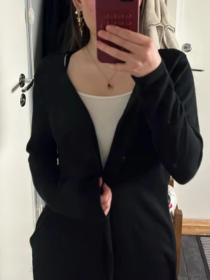 Svart lång kofta i mjukt material - Säljer en svart lång kofta från h&m med enkel design och långa ärmar. Koftan är öppen framtill och gjord i ett mjukt, tunt material. Storlek S