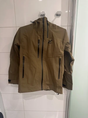 Grön vindjacka RevolutionRace S - Säljer en grön vindjacka från RevolutionRace cyclone 3L shell i färgen dark olive, dammodell, storlek S. Jackan har flera dragkedjeförsedda fickor, huva och RECCO-reflektor. Materialet är slitstark polyester och insidan har meshficka. Perfekt för uteliv och äventyr. I fint skick, nypris 2000 kr