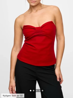 Röd bandeau topp med rosett från Bik Bok - Säljer en röd bandeau topp från Bik Bok (aldrig använd) med en stor rosett framtill. Toppen är axelbandslös men har tunna avtagbara band och dragkedja i ryggen. Materialet är mjukt och lätt, perfekt för att sticka ut. Supersnygg till både jeans och kjol! 