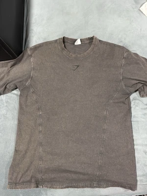 Mörkgrå Gymshark t-shirt S - Säljer en mörkgrå overseized t-shirt från Gymshark i storlek S. T-shirten har långa armar , rund halsringning och en diskret Gymshark-logga framtill samt stor text på ryggen. Tillverkad i mjuk bomull, perfekt för gymmet eller chill