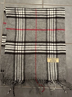 Grå rutig Burberry väst i kashmir - Säljer en klassisk grå väst från Burberry i 100% kashmir. Västen har ett rutigt mönster i svart, vitt och rött samt fransar nedtill. Märkeslapp med Burberry London på framsidan. Perfekt för dig som vill ha en tidlös och lyxig look.