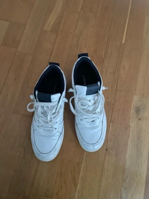 Vita Björn Borg sneakers med svarta detaljer - Säljer ett par vita sneakers från Björn Borg med mörkblå detaljer och logga på hälen. Skorna har snörning, platt sula och rund tå. Tillverkade i syntetmaterial och har en klassisk, stilren look som passar till allt en sko för komplimanger och komfort. Pris går att diskutera 