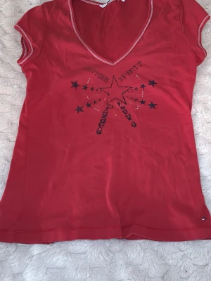 Röd Tommy Hilfiger t-shirt med stjärntryck - Röd t-shirt från Tommy Hilfiger med coolt stjärntryck och texten 'Hilfiger Spirit' framtill. T-shirten har v-ringning, korta ärmar och kontrastsömmar vid hals och ärmslut. Tillverkad i mjuk bomull som känns skön mot huden. Storleken är L men jag kan använda den och jag brukar ha xs 