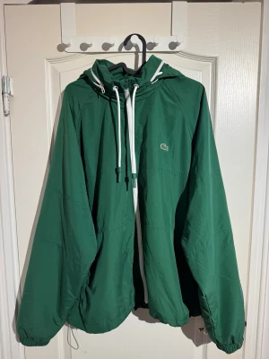 Grön vindjacka från Lacoste L/XL - Säljer en grön vindjacka från Lacoste i storlek L/XL. Jackan har huva med vita snören, vit dragkedja framtill och det klassiska Lacoste-märket på bröstet. Tillverkad i lätt polyester med meshfoder för extra komfort. Perfekt för dig som gillar sportig och clean stil. Vid minsta fråga, hör av dig 🙌🏽