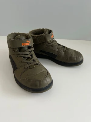 Olivgröna sneakers från Puma - Snygga olivgröna sneakers från Puma med svart detalj på sidan och orange logga. Skorna har snörning, kardborreband och är fodrade med mjukt material för extra värme. Perfekta för kyligare dagar och har en klassisk rund tå och platt sula.
