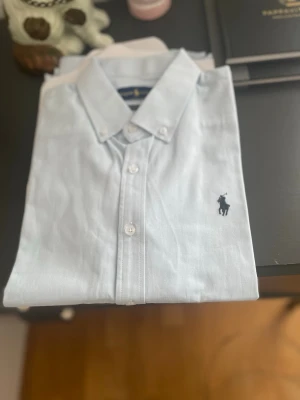 Ljusblå Ralph Lauren skjorta - Klassisk ljusblå skjorta från Ralph Lauren med button-down krage och broderad logga på bröstet. Skjortan har lång ärm, vita knappar och är i en skön bomullskvalitet. Perfekt till jeans eller chinos för en clean look.