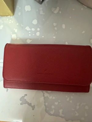 Röd plånbok från Daniel Hechter - Säljer en stilren röd plånbok från Daniel Hechter i skinn. Plånboken har flera fack, ett innerfack med dragkedja och stängs med tryckknapp. Perfekt storlek för att hålla koll på kort, kontanter och smågrejer. Nästan oanvänd 