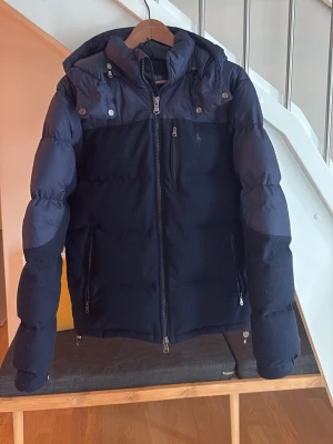 Mörkblå pufferjacka från Polo Ralph Lauren - Säljer en mörkblå pufferjacka från Polo Ralph Lauren med huva och dragkedjor framtill.  Jackan är köpt föra året från zalando och använd 1 halv vinter, nypris 6000, mitt pris 3000 