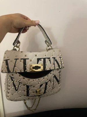  Valentino Rockstud väska  - Säljer en superfin vit handväska från Valentino med stora svarta VLTN-bokstäver och guldiga nitar. Väskan har quiltad design, guldfärgade detaljer och kedjerem. Insidan är röd och har dragkedja. Perfekt för dig som vill sticka ut med en lyxig accessoar.
