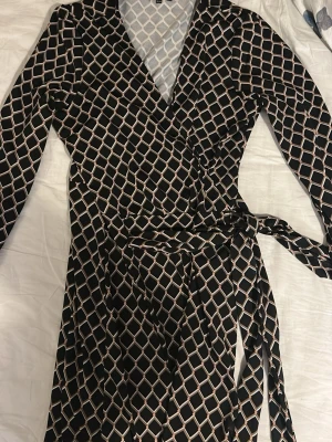 Mönstrad omlottklänning från Happy Holly - Säljer en svart omlottklänning från Happy Holly med långa ärmar och knytband i midjan. Klänningen har ett rutigt mönster i beige och ljusrosa toner, vilket ger en snygg och modern look. Perfekt för dig som gillar stilrena detaljer och bekväm passform.