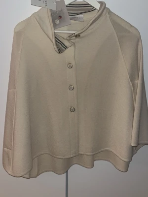 Beige väst från Busnel med knappar - Snygg beige väst från Busnel med rund hals och dekorativa guldknappar framtill. Västen har en mjuk, stickad känsla och en avslappnad passform som faller snyggt över axlarna. Perfekt att slänga över en tröja för en stilren look.