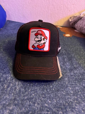 Super Mario keps från Capslab - Säljer en svart och röd keps från Capslab med Super Mario-motiv framtill. Kepsen har mesh på sidorna och bak, röda sömmar och justerbar snapback. Framsidan är i tyg och skärmen är böjd. Perfekt för dig som gillar gaming och streetstyle.