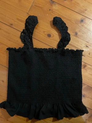 Svart volangtop från New Look - Supersöt svart topp från New Look med breda axelband i broderad stil och volangkant nertill. Smockad över hela kroppen för en bekväm och stretchig passform. Perfekt till jeans eller kjol 