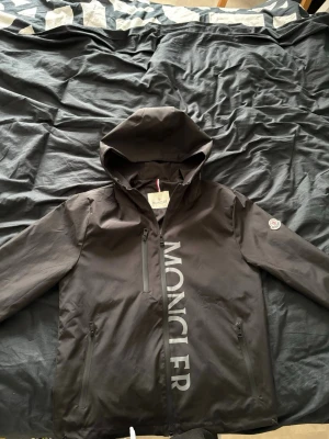 Svart vindjacka från Moncler - Säljer en svart vindjacka från Moncler med stor logga längs dragkedjan och klassisk Moncler-patch på ärmen. Jackan har huva, tre fickor med dragkedja och en clean, sportig look. Materialet är syntet och jackan är perfekt för blåsiga dagar.