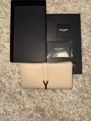 Beige plånbok från Saint Laurent - Säljer en lyxig beige plånbok från Saint Laurent Paris i skinn med guldiga detaljer och dragkedja. Plånboken har flera kortfack och ett stort Y-spänne framtill. Kommer med originalbox och även med certifikat att den är äkta. Den är helt oanvänd. Nypriset på denna vara e runt 5000kr. Kontakta via meddelande för att se certifikat