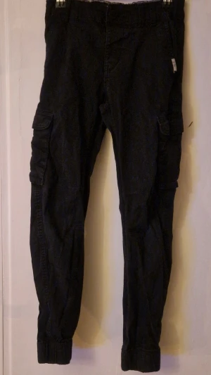 Svarta cargobyxor från KappAhl  - Säljer ett par svarta cargopants i storlek 140 från KappAhl med elastisk midja och muddar vid bensluten. Byxorna har flera praktiska fickor på sidorna och bak, samt ställbar midja. Aldrig använda. Hund finns i hemmet. Kolla in mina andra annonser för samfrakt! 