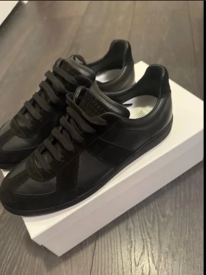 Svarta Maison Margiela sneakers - Säljer ett par svarta sneakers från Maison Margiela med klassisk design. Skorna har en mix av mocka och skinn, svarta snören och en låg siluett. Perfekta för dig som gillar stilrena och tidlösa skor med exklusiv känsla. Vet ej om det är ekta så pris kan diskuteras vid köp, det är helt nya och aldrig använda, säljer det på grund av fel storlek. 