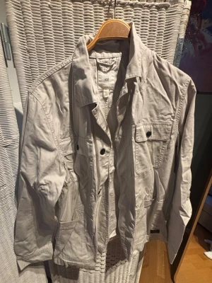 Beige utilityjacka från H&M - Stilren beige utilityjacka från H&M med flera praktiska fickor framtill och knappar. Jackan har en avslappnad passform och klassisk krage, perfekt för lager på lager. Tillverkad i bomull och passar till många olika looks.