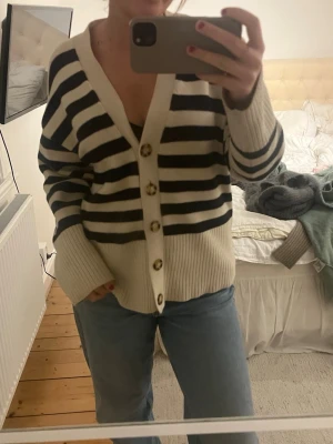 Randig beige och blå kofta - Säljer en oversized kofta med breda mörkblå och beige ränder. Koftan har djup v-ringning, stora knappar framtill och ribbade muddar vid ärmsluten. Perfekt att slänga över en t-shirt för en avslappnad look. 