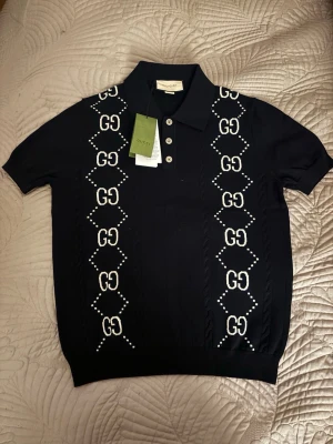 Svart kortärmad topp från Gucci - Snygg svart topp från Gucci med korta ärmar och klassisk krage. Framsidan har vita GG-loggor i ett mönster längs båda sidorna och knappar framtill. Materialet känns mjukt och strechigt 