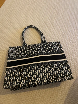 Svartvit shoppingväska med mönster - Säljer en snygg svartvit shoppingväska med grafiskt mönster och två handtag. Väskan har en rektangulär form och är tillverkad i tyg, perfekt för att bära med sig allt man behöver. Den har ett coolt, upprepande mönster och breda ränder som ger den en trendig look.