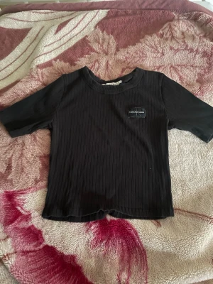 Svart ribbad topp Calvin Klein - Säljer en svart ribbad topp från Calvin Klein med korta ärmar och rund hals. Toppen har en liten logotyp på bröstet och är croppad i modellen. Perfekt till jeans eller kjol för en trendig look.