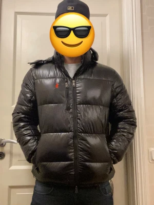 Polo Ralph Lauren jacka  - Snygg svart pufferjacka från Polo Ralph Lauren med blank finish och röd logga på bröstet. Jackan har huva, flera dragkedjefickor och quiltad design. Perfekt för kalla dagar och ger en modern och varm look. 