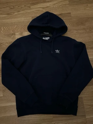 Mörkblå hoodie från Adidas - Snygg mörkblå hoodie från Adidas med klassisk vit logga på bröstet. Hoodien har huva med dragsko och en stor magficka framtill. Tillverkad i mjukt material som känns skönt mot huden. Perfekt för en avslappnad och sportig stil.