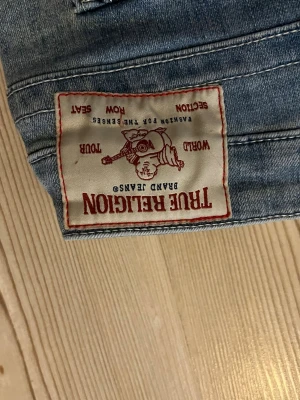 Blå jeans från True Religion - Säljer ett par klassiska blå jeans från True Religion med ikonisk patch bak och raka ben. Jeansen har tydliga sömmar och en ficka med knappdetalj bak. Perfekta för dig som gillar streetwear och vill ha en avslappnad look.