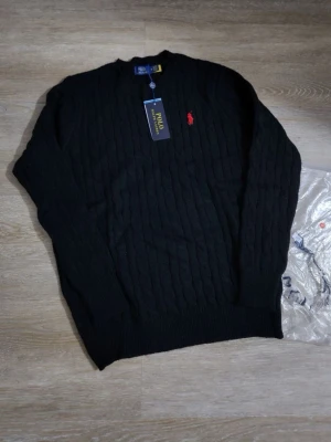 Svart kabelstickad tröja Polo Ralph Lauren - Kabelstickad svart tröja från Polo Ralph Lauren med klassisk röd logga broderad på bröstet. Tröjan har rund halsringning och ribbade muddar vid ärmslut och nederkant. Perfekt för en stilren och tidlös look.