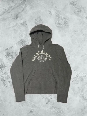 Grå hoodie från Abercrombie - Mörkgrå hoodie från Abercrombie med vit logga och text på bröstet. Klassisk känguruficka framtill, justerbar dragsko i huvan och ribbade muddar. Perfekt för en avslappnad och stilren look. Storleken är s. Inga defekter. Tveka inte att skriva om du undrar något.