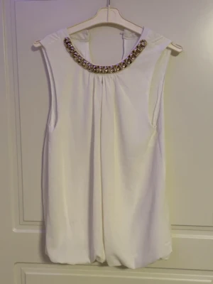 Vit ärmlös blus med strassdetaljer - Stilren vit blus i ett lätt, flödande tyg. Rund halsringning dekorerad med gnistrande strass och pärlor som ger en lyxig touch. Blusen är ärmlös och har en lös passform med diskret knappstängning i nacken. Perfekt för att lyfta vilken outfit som helst.