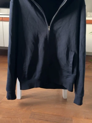 Svart ziptröja med fickor - En stilren svart ziptröja med hel dragkedja framtill och två sidofickor. Tröjan har långa ärmar och ribbade muddar vid ärmslut och nederkant. Perfekt för en avslappnad look och enkel att matcha med det mesta.