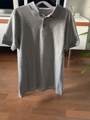 Grå pikétröja i bomull - Säljer en stilren grå pikétröja med klassisk krage och knappar framtill. Tröjan har korta ärmar och är gjord i mjukt bomullsmaterial. 