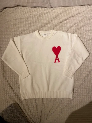 Vit stickad tröja från Ami Paris - Säljer en vit stickad tröja från Ami Paris med deras ikoniska röda hjärta och bokstaven A broderat på bröstet. Tröjan har rund hals, långa ärmar och ribbade muddar vid ärmslut och nederkant. Perfekt för dig som gillar stilrena och trendiga plagg. Bara att höra av dig vid frågor😊 