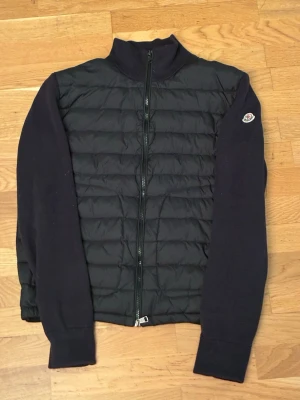Svart dunväst från Moncler - Snygg svart dunväst från Moncler med quiltad front och hel dragkedja. Västen har ribbstickade ärmar och Moncler-logga på ena ärmen. Perfekt för lager-på-lager och streetstyle. Materialet är en mix av dun och stickad bomull.