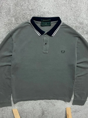 Grå långärmad piké från Fred Perry - Snygg grå långärmad pikétröja från Fred Perry med klassisk svart krage och vita ränder. Tröjan har knappar vid halsen och den ikoniska Fred Perry-loggan broderad på bröstet. Perfekt för en clean och stilren look.