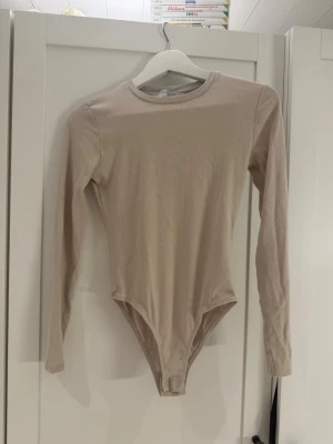 Beige långärmad body Primark XS - Stilren beige body från Primark i storlek XS. Modellen har långa ärmar, rund halsringning och knäppning i grenen. Tillverkad i mjukt, stretchigt material som sitter skönt mot kroppen. Perfekt att matcha med jeans eller kjol för en enkel och clean look.