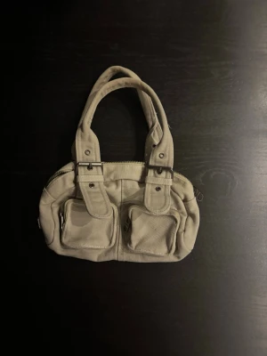 Beige handväska med dubbla fickor - Snygg beige handväska i canvas med två framfickor med dragkedja och dubbla handtag. Väskan har en enkel, stilren design och metallspännen på handtagen för en cool touch. Perfekt för dig som gillar praktiska detaljer och minimalistisk stil.