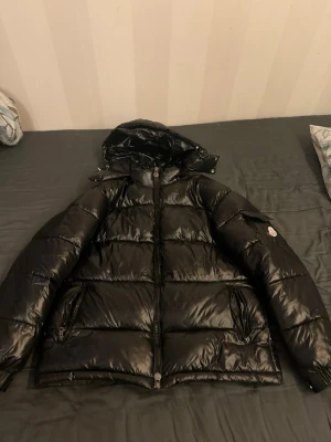 Svart Moncler dunjacka med huva - Snygg svart dunjacka från Moncler med glansig finish och huva. Jackan har dragkedja framtill, två fickor med dragkedja och en ficka med knapp på ärmen där Moncler-loggan sitter. Perfekt för kalla dagar och riktigt trendig look.