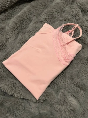 Ljusrosa linne med spetsdetalj - Sött ljusrosa linne med tunna axelband och v-ringning dekorerad med spets. Perfekt för mysiga hemmakvällar eller som loungewear. Mjukt material som känns skönt mot huden.