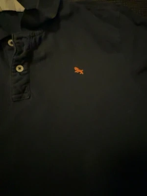 Mörkblå piké med orange logga - Mörkblå pikétröja med klassisk krage och två knappar framtill. På bröstet finns en liten orange broderad logga. Skjortan har en enkel och stilren design, perfekt för en avslappnad look.