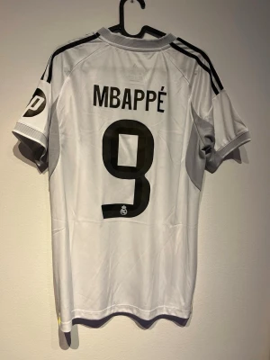 Real Madrid Mbappé matchtröja vit - Snygg vit Real Madrid matchtröja från Adidas med Mbappé och nummer 9 på ryggen. Tröjan har svarta detaljer, klubbmärke på bröstet och sponsortryck framtill. Tillverkad i lätt och ventilerande material med AEROREADY-teknologi. Perfekt för fotbollsträning eller supporterliv.