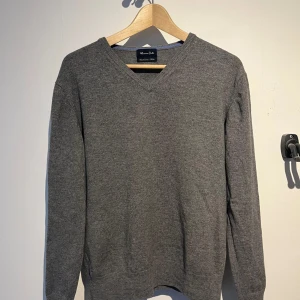 Grå tröja från Massimo Dutti - Säljer en stilren grå V-neck tröja från Massimo Dutti. Tröjan har en v-ringad design och är tillverkad i Cashmere. Den är i bra skick förutom ett mindre hål på högra armen, därav priset. Tröjan passar ca. 175. Hör av er vid frågor!