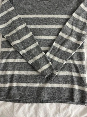 Gråvit randig stickad tröja - Mysig stickad tröja i grått med vita horisontella ränder. Tröjan har rund halsringning och långa ärmar. Perfekt för lager på lager och chill dagar. Materialet känns mjukt och bekvämt mot huden.
