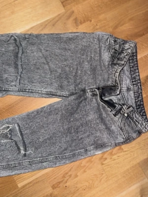 Grå tvättade jeans med slitning - Säljer ett par grå tvättade jeans med rak passform och en snygg slitning på ena benet. Jeansen har klassisk femficksdesign och normal midja. Perfekta för dig som gillar en avslappnad och trendig look.