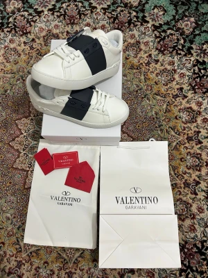 Valentino Garavani vita sneakers - Säljer ett par vita Valentino Garavani sneakers med bred svart rem över vristen och guldfärgad logga på plösen. Skorna har klassisk rund tå, snörning och platt sula. Kommer med originalkartong, dustbag och tags.