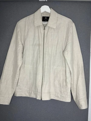 Beige jacka i linne från Cavour - Stilren beige jacka från Cavour i linne med rak passform och klassisk krage. Jackan har dragkedja framtill, två sidofickor och långa ärmar. Perfekt för vår och sommar när du vill ha en lätt och luftig jacka.